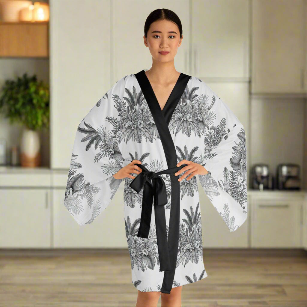 Woman wearing a chic long sleeve kimono robe with black and white tropical floral print and black trim, tied at the waist, standing in a modern kitchen.