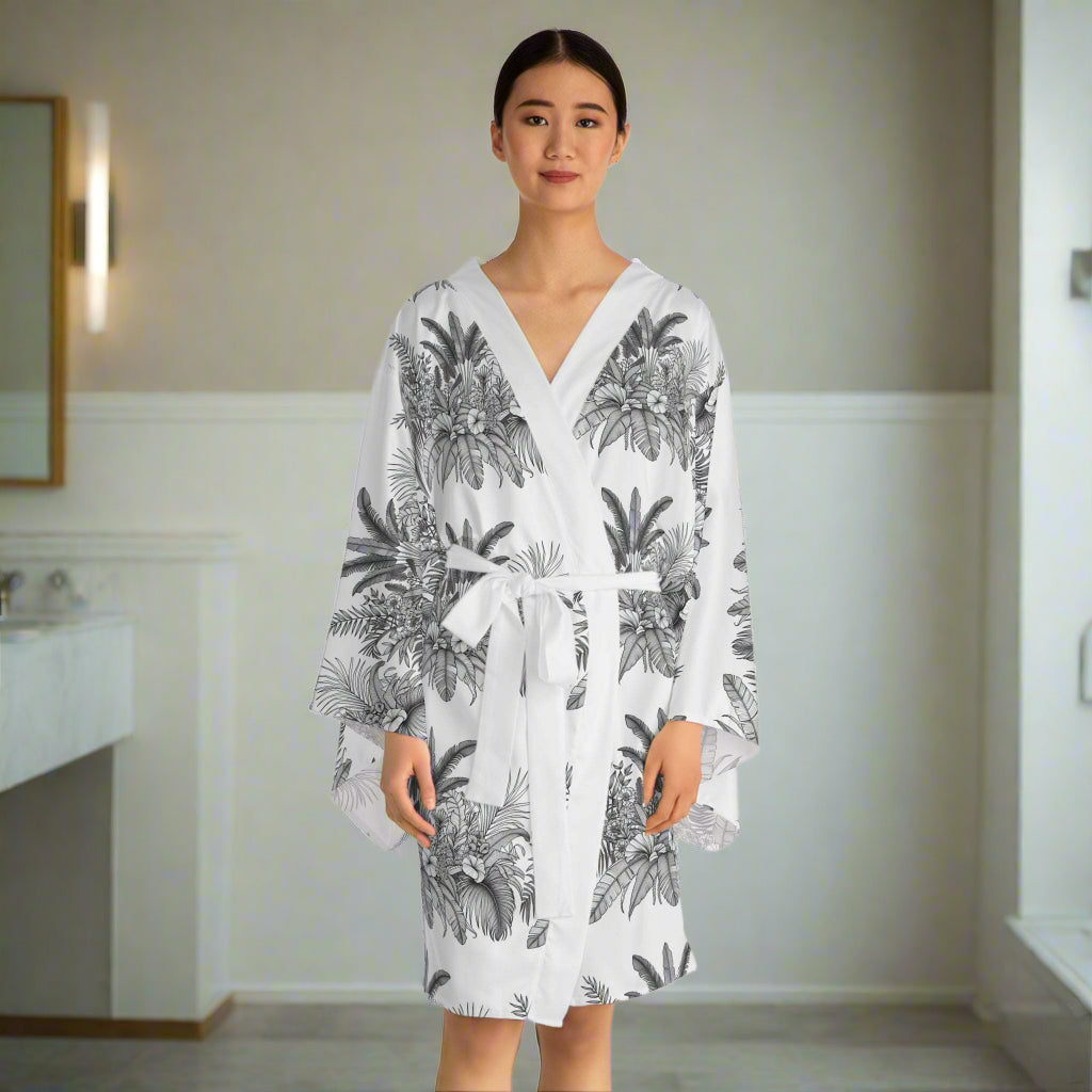 Woman wearing a chic white floral long sleeve kimono robe with tropical black leaf print and a waist tie belt.